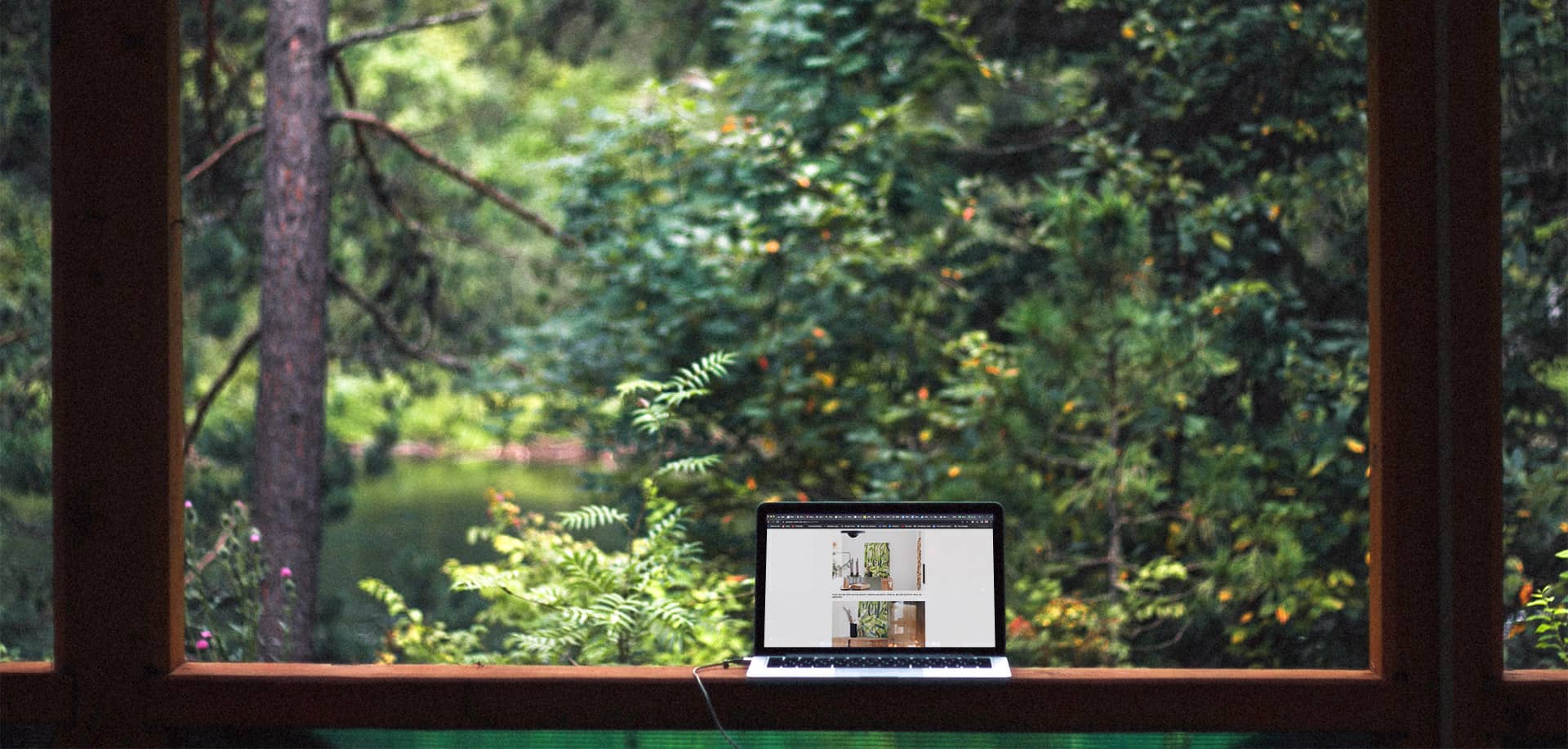 Laptop in einer Lodge auf einer Fensterbank, Blick in ein sommerliches Waldstück mit Laub- und Nadelbäumen und einem Weiher